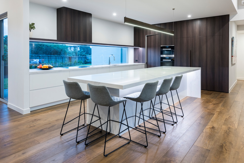 Inspiration for a contemporary l-shaped kitchen in Melbourne with a drop-in sink, flat-panel cabinets, quartz benchtops, window splashback, stainless steel appliances, medium hardwood floors, multiple islands, brown floor and white benchtop.