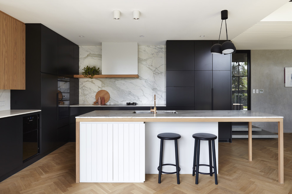 This is an example of a contemporary l-shaped open plan kitchen in Adelaide with an undermount sink, black cabinets, marble benchtops, white splashback, marble splashback, black appliances, light hardwood floors, with island and white benchtop.