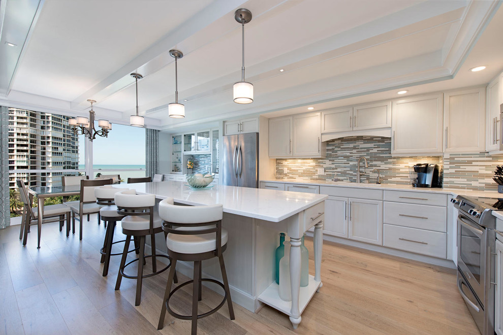 This is an example of a large beach style l-shaped open plan kitchen in Tampa with an undermount sink, white cabinets, quartz benchtops, multi-coloured splashback, glass tile splashback, stainless steel appliances, light hardwood floors, with island, shaker cabinets and beige floor.
