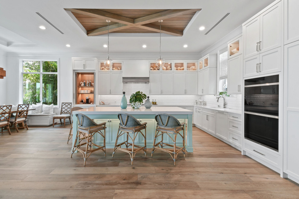 This is an example of a medium sized nautical l-shaped kitchen in Other with white cabinets, engineered stone countertops, white splashback, metro tiled splashback, integrated appliances, light hardwood flooring, an island, brown floors, white worktops and a drop ceiling.