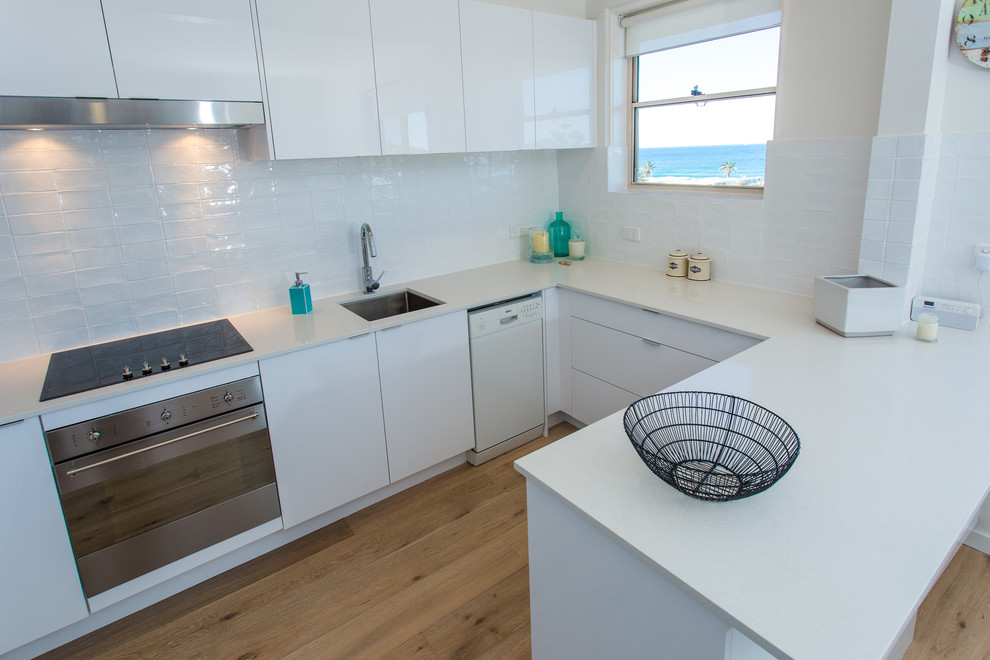 This is an example of a small beach style u-shaped eat-in kitchen in Sydney with a drop-in sink, flat-panel cabinets, white cabinets, quartz benchtops, white splashback, subway tile splashback, stainless steel appliances, light hardwood floors and no island.