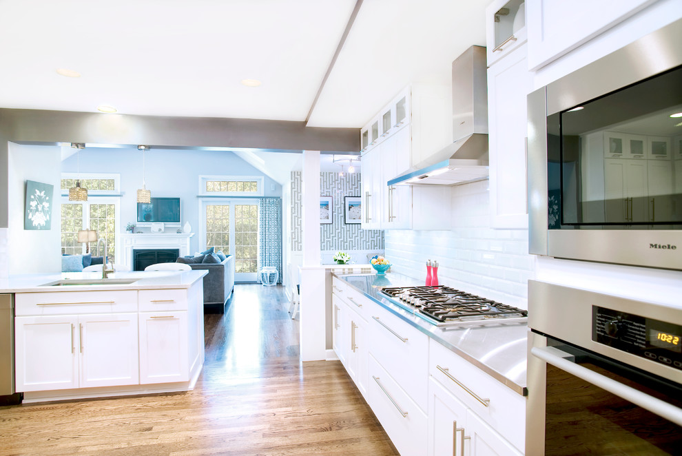 Inspiration for a mid-sized transitional l-shaped eat-in kitchen in Chicago with an undermount sink, shaker cabinets, white cabinets, stainless steel benchtops, white splashback, subway tile splashback, stainless steel appliances, medium hardwood floors and a peninsula.
