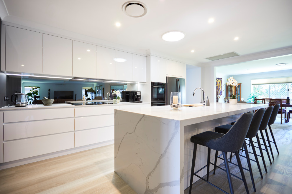 Design ideas for a large modern galley eat-in kitchen in Sunshine Coast with recessed-panel cabinets, white cabinets, quartz benchtops, metallic splashback, mirror splashback, stainless steel appliances, with island, white benchtop, an integrated sink, light hardwood floors and beige floor.