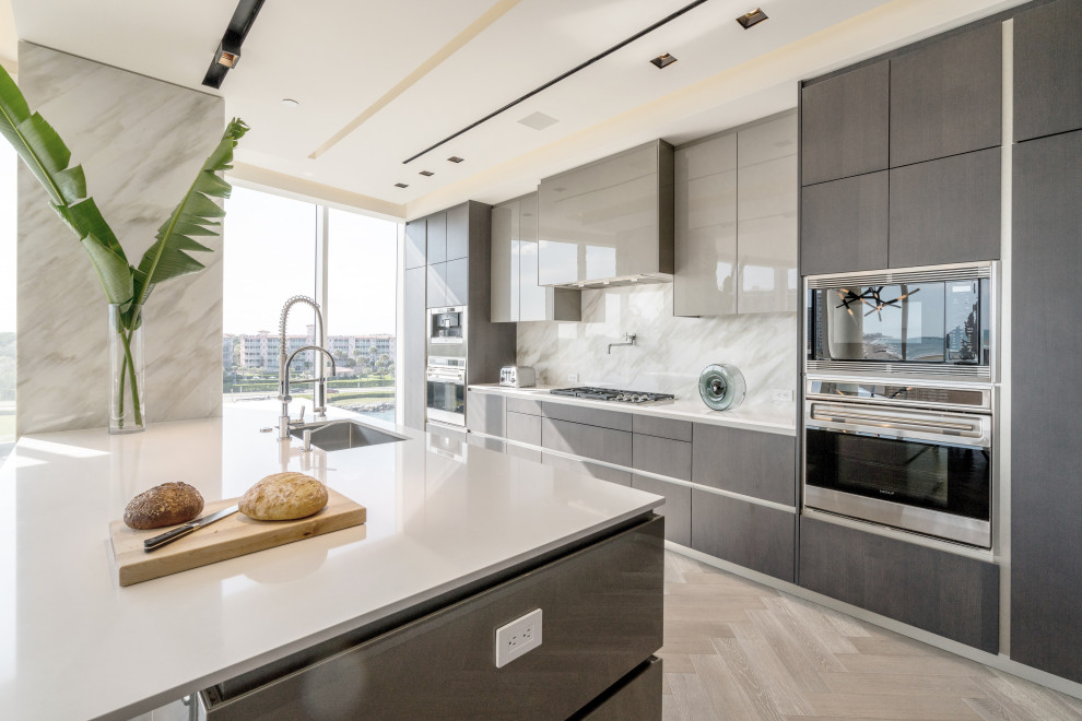 Photo of a contemporary galley kitchen in Miami with an undermount sink, flat-panel cabinets, grey cabinets, grey splashback, stone slab splashback, stainless steel appliances, light hardwood floors, with island, beige floor and white benchtop.