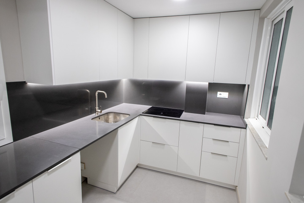 Bild på ett avskilt, mellanstort funkis grå grått l-kök, med en enkel diskho, släta luckor, vita skåp, grått stänkskydd, svarta vitvaror, klinkergolv i keramik och grått golv