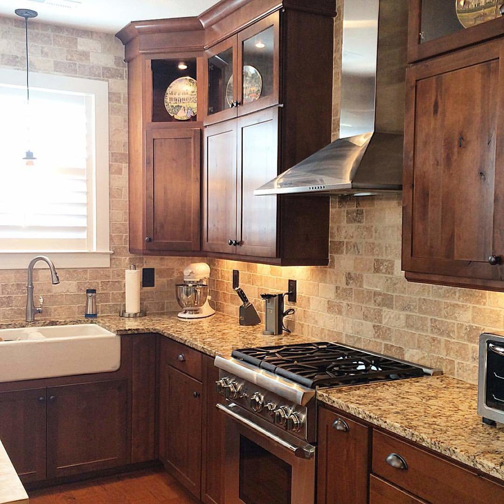 Large transitional l-shaped light wood floor eat-in kitchen photo with a farmhouse sink, shaker cabinets, brown cabinets, granite countertops, beige backsplash, ceramic backsplash, stainless steel appliances and an island