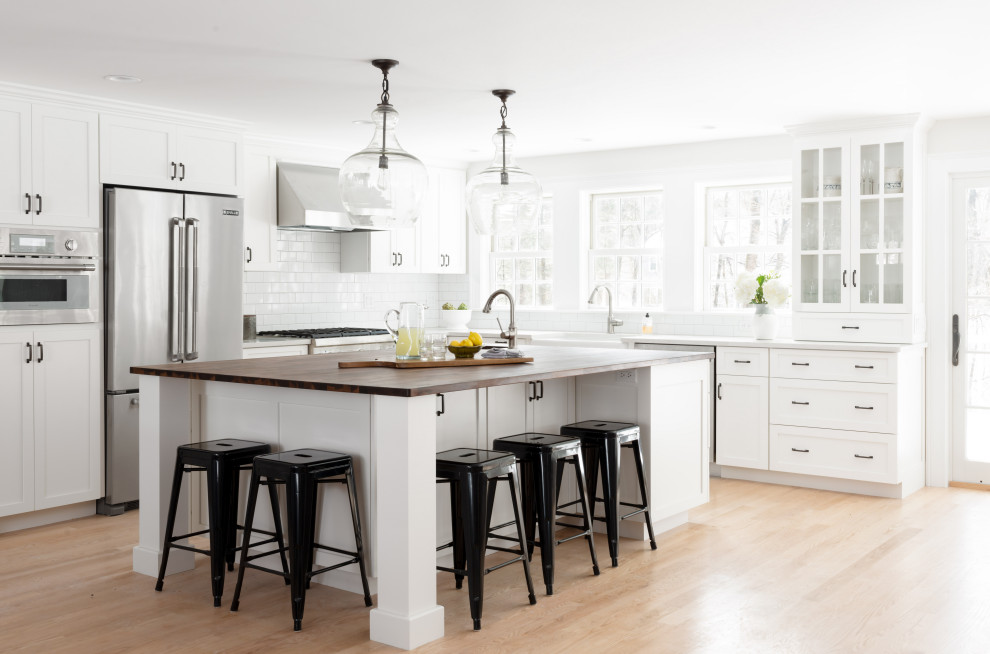 Example of a transitional l-shaped light wood floor and beige floor kitchen design in Boston with a farmhouse sink, shaker cabinets, white cabinets, white backsplash, subway tile backsplash, stainless steel appliances, an island and white countertops