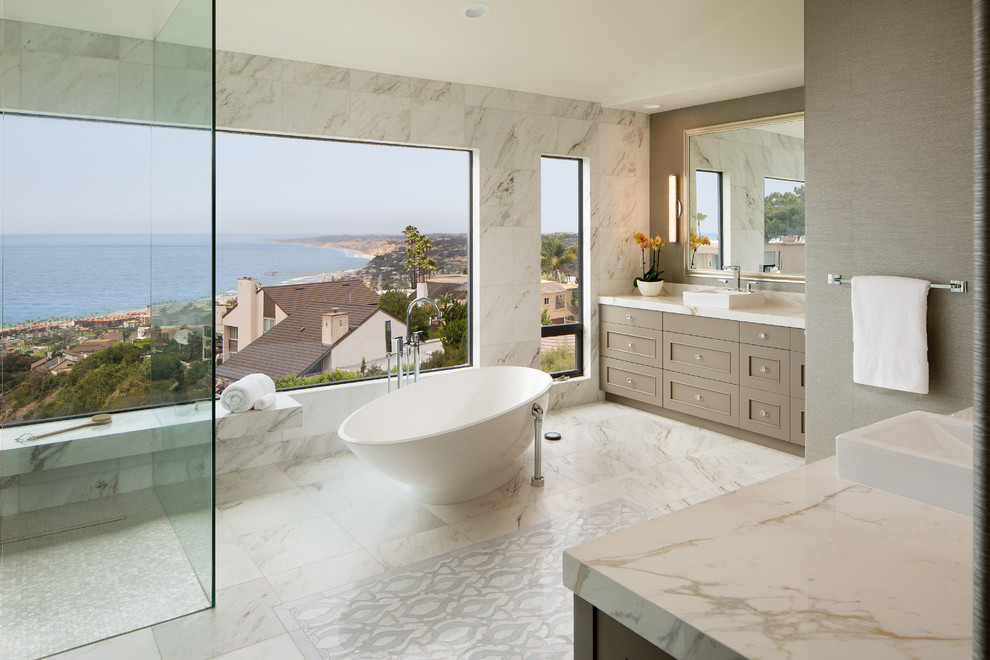 This is an example of an expansive beach style master bathroom in San Diego with shaker cabinets, beige cabinets, a freestanding tub, a curbless shower, a two-piece toilet, black and white tile, marble, grey walls, marble floors, a vessel sink, marble benchtops, blue floor, a hinged shower door and white benchtops.