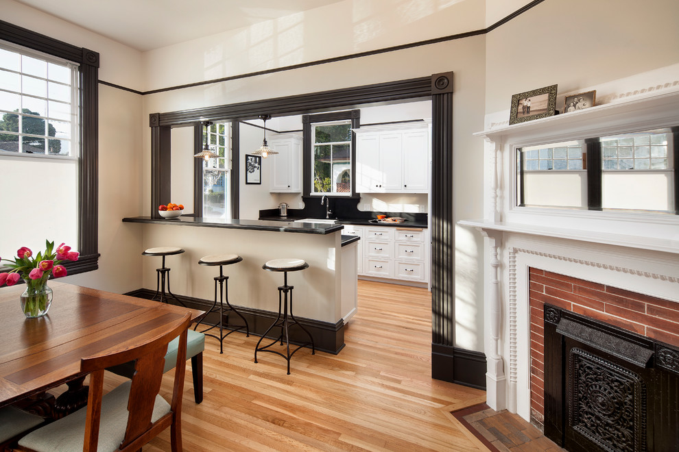 This is an example of a small traditional galley eat-in kitchen in Santa Barbara with recessed-panel cabinets, white cabinets, granite benchtops, black splashback, light hardwood floors, stainless steel appliances and a farmhouse sink.