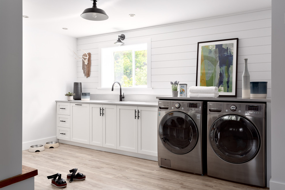 Design ideas for a mid-sized country single-wall utility room in Vancouver with an undermount sink, shaker cabinets, white cabinets, white walls, light hardwood floors, a side-by-side washer and dryer, beige floor, grey benchtop and planked wall panelling.