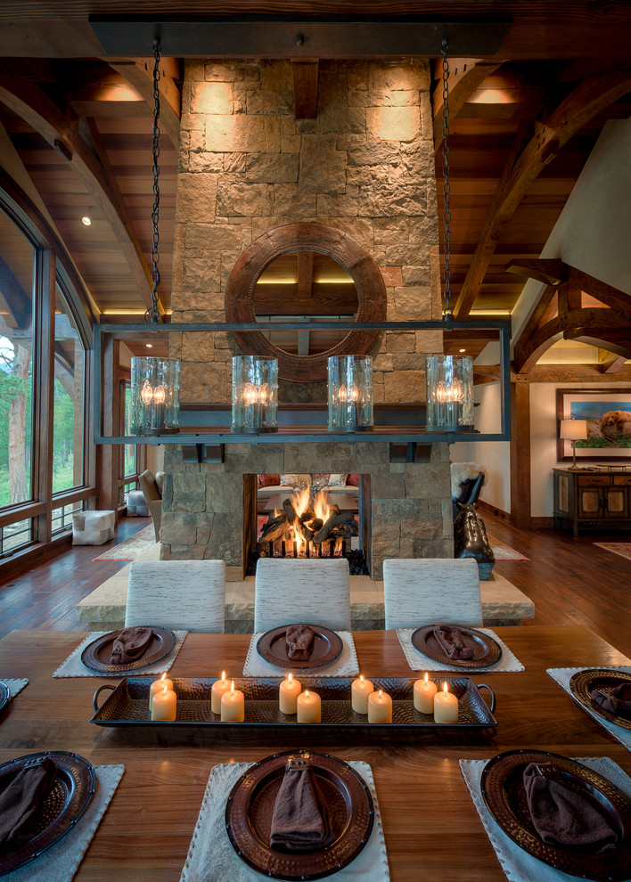 This is an example of a large country open plan dining in Denver with beige walls, dark hardwood floors, a two-sided fireplace, a stone fireplace surround and brown floor.