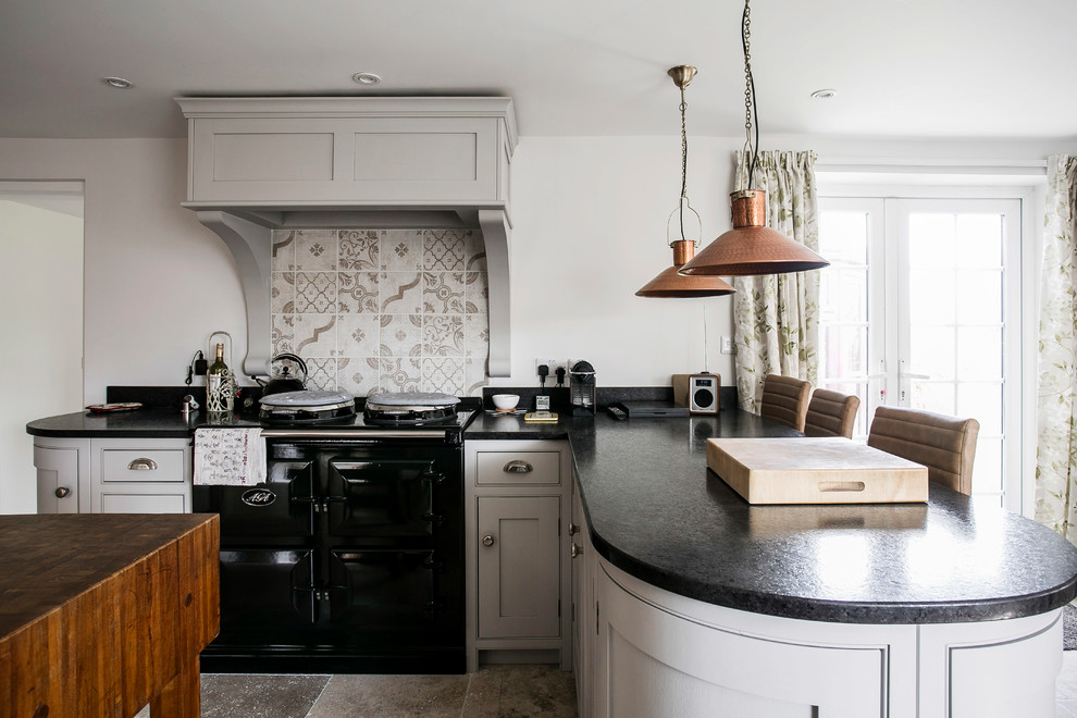 Design ideas for a mid-sized traditional u-shaped open plan kitchen in Kent with a farmhouse sink, shaker cabinets, grey cabinets, granite benchtops, brown splashback, ceramic splashback, black appliances, limestone floors, with island, beige floor and black benchtop.