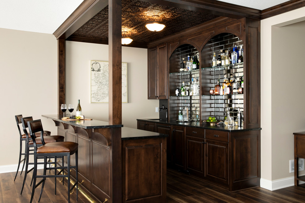 This is an example of a mid-sized transitional galley seated home bar in Minneapolis with an undermount sink, raised-panel cabinets, dark wood cabinets, soapstone benchtops, mirror splashback, vinyl floors, brown floor and black benchtop.