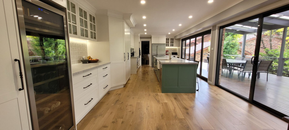 This is an example of a large l-shaped open plan kitchen in Sydney with a farmhouse sink, shaker cabinets, green cabinets, quartz benchtops, grey splashback, porcelain splashback, stainless steel appliances, medium hardwood floors, with island, brown floor, multi-coloured benchtop and recessed.