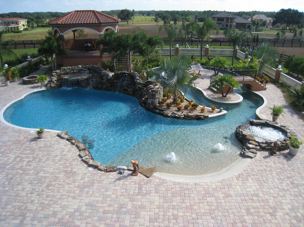 Photo of an expansive tropical backyard custom-shaped pool in Tampa with a water feature and brick pavers.