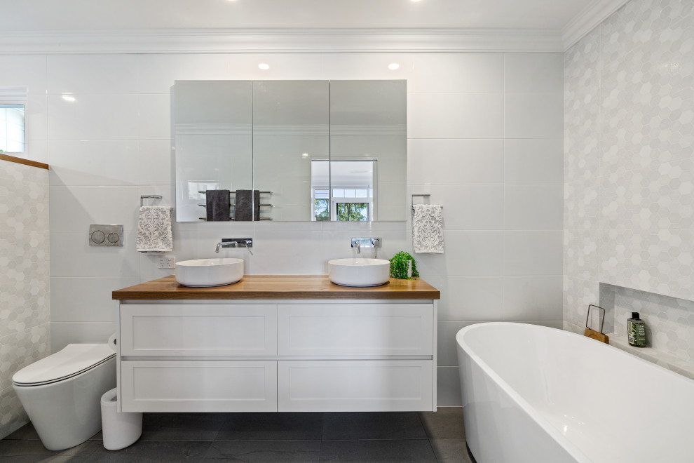 This is an example of a beach style bathroom in Central Coast with shaker cabinets, white cabinets, a freestanding tub, white tile, a vessel sink, wood benchtops, brown benchtops, a double vanity, a floating vanity, a one-piece toilet and grey floor.
