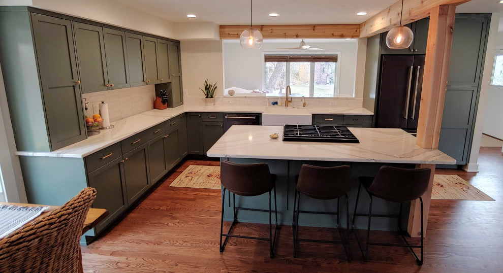 Example of a mid-sized transitional l-shaped dark wood floor eat-in kitchen design with a farmhouse sink, recessed-panel cabinets, green cabinets, quartz countertops, white backsplash, black appliances, an island and white countertops