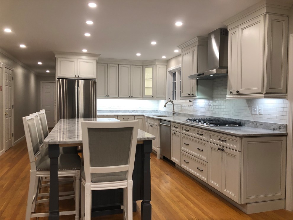 Example of a large transitional l-shaped medium tone wood floor and brown floor open concept kitchen design in Boston with an undermount sink, recessed-panel cabinets, white cabinets, quartz countertops, white backsplash, subway tile backsplash, stainless steel appliances, an island and multicolored countertops