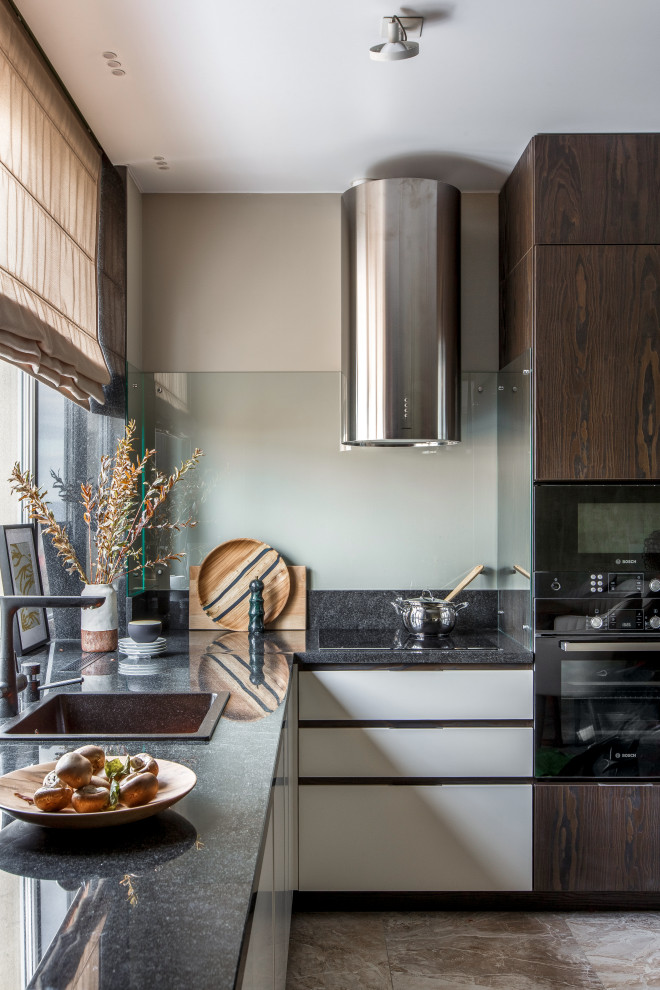 Mid-sized contemporary l-shaped kitchen in Other with a drop-in sink, flat-panel cabinets, white cabinets, glass sheet splashback, black appliances, no island, beige floor and black benchtop.