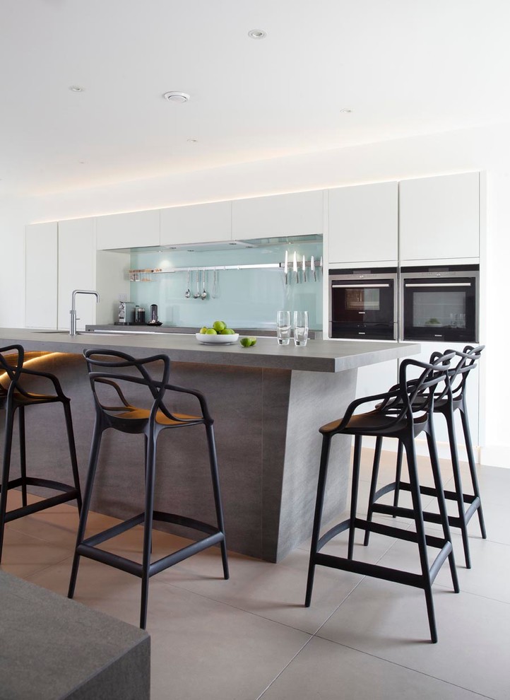 Expansive contemporary u-shaped open plan kitchen with an integrated sink, flat-panel cabinets, white cabinets, engineered stone countertops, green splashback, glass sheet splashback, stainless steel appliances, ceramic flooring and an island.