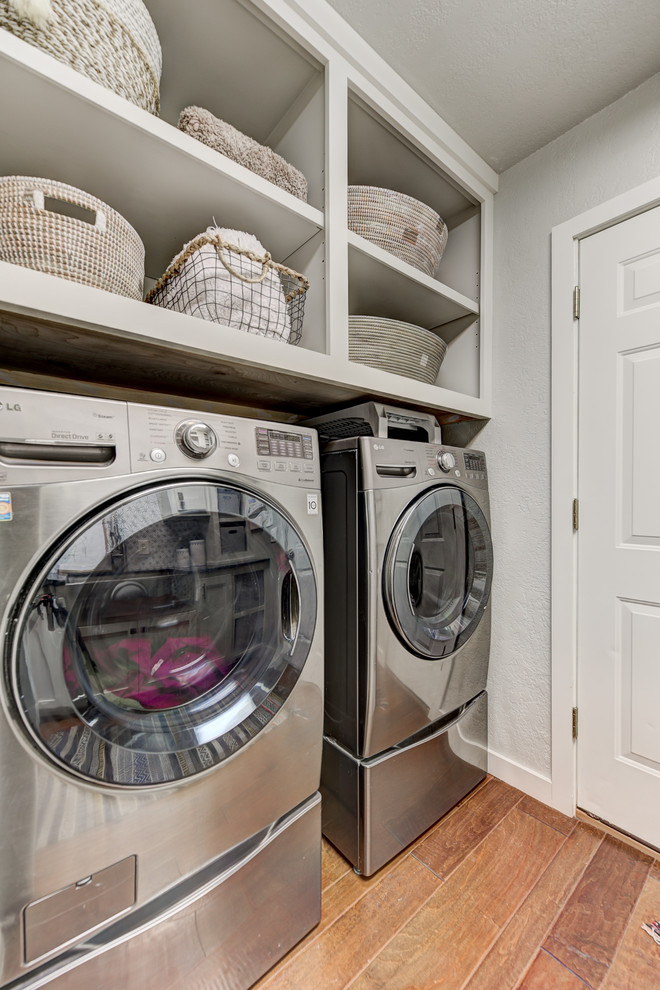 Design ideas for a mid-sized transitional galley dedicated laundry room in Oklahoma City with a farmhouse sink, shaker cabinets, white cabinets, granite benchtops, grey walls, medium hardwood floors, a side-by-side washer and dryer, brown floor and black benchtop.