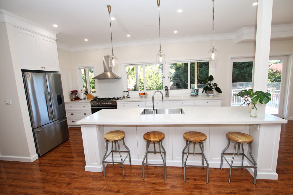 This is an example of a large transitional galley open plan kitchen in Sydney with a double-bowl sink, shaker cabinets, white cabinets, quartz benchtops, white splashback, subway tile splashback, stainless steel appliances, medium hardwood floors, with island, brown floor and white benchtop.