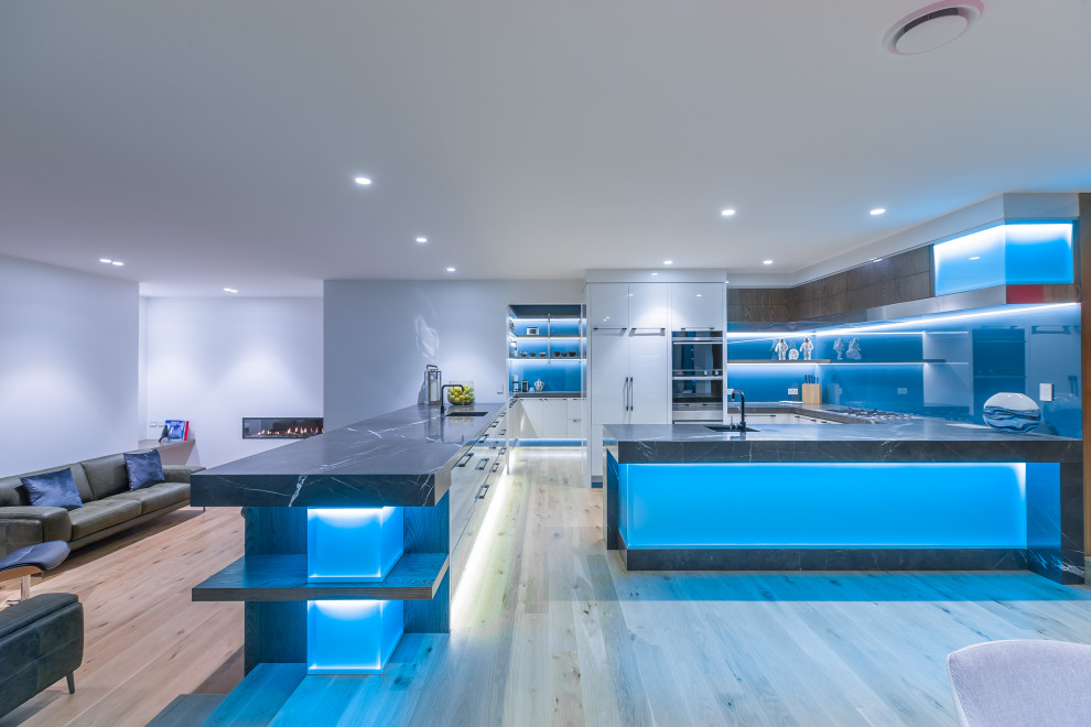 Expansive contemporary l-shaped open plan kitchen in Auckland with a submerged sink, flat-panel cabinets, white cabinets, granite worktops, blue splashback, glass sheet splashback, stainless steel appliances, light hardwood flooring, multiple islands, brown floors and grey worktops.
