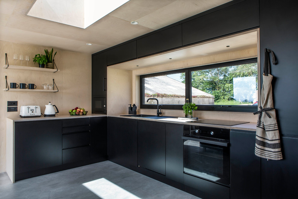 This is an example of a scandinavian l-shaped kitchen in Oxfordshire with a drop-in sink, flat-panel cabinets, black cabinets, wood benchtops, window splashback, black appliances, concrete floors, grey floor and beige benchtop.