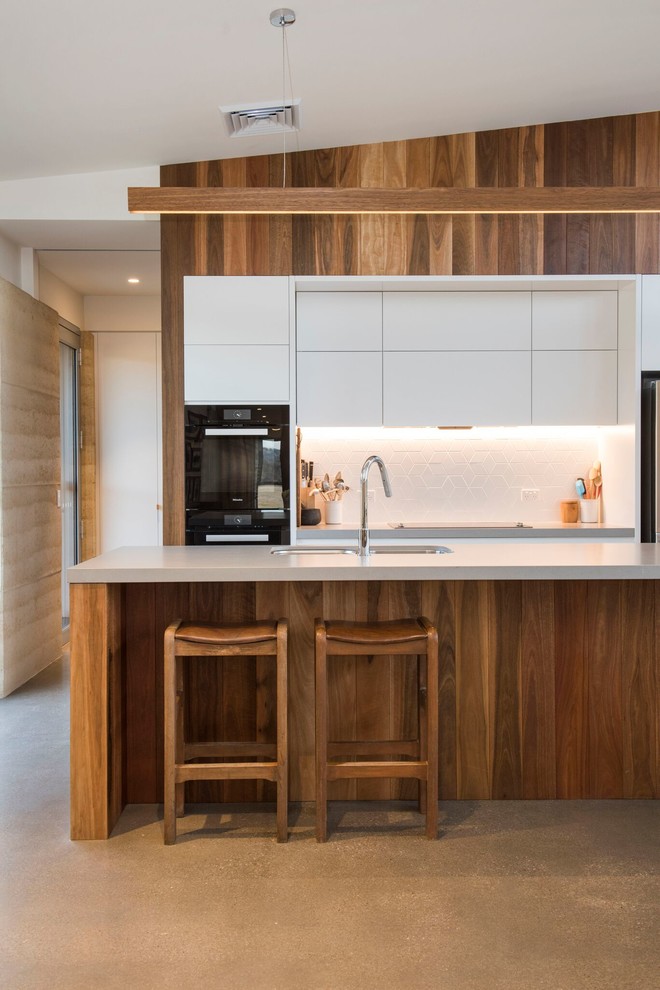 Photo of a contemporary galley kitchen in Other with a double-bowl sink, flat-panel cabinets, white cabinets, white splashback, black appliances, concrete floors, with island, grey floor and grey benchtop.