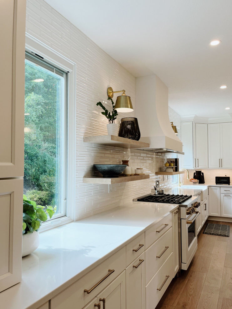 Eat-in kitchen - large transitional l-shaped light wood floor and beige floor eat-in kitchen idea with a farmhouse sink, shaker cabinets, white cabinets, quartz countertops, white backsplash, ceramic backsplash, white appliances, an island and white countertops