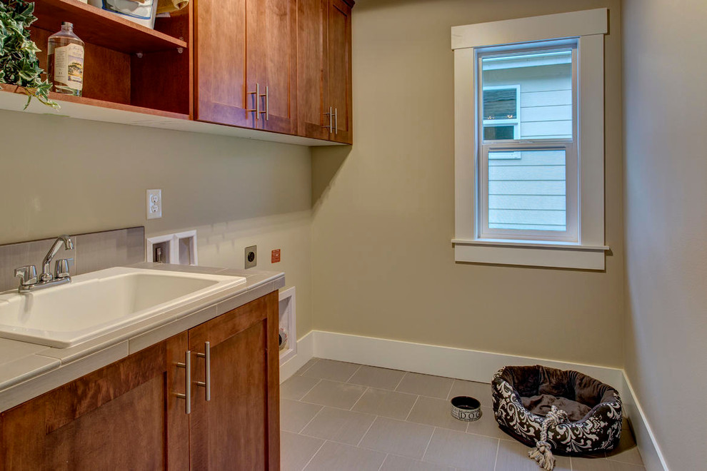 This is an example of a mid-sized single-wall utility room in Seattle with open cabinets, medium wood cabinets, beige walls, porcelain floors, a drop-in sink, tile benchtops and a side-by-side washer and dryer.