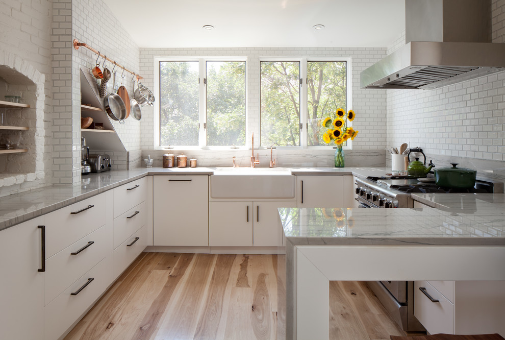 Inspiration for a contemporary u-shaped eat-in kitchen in Boston with a farmhouse sink, flat-panel cabinets, white cabinets, white splashback, stainless steel appliances, light hardwood floors, a peninsula and subway tile splashback.