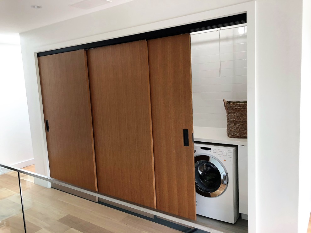 Small modern single-wall laundry cupboard in Toronto with an undermount sink, quartz benchtops, white walls, a side-by-side washer and dryer, white benchtop, light hardwood floors and brown floor.