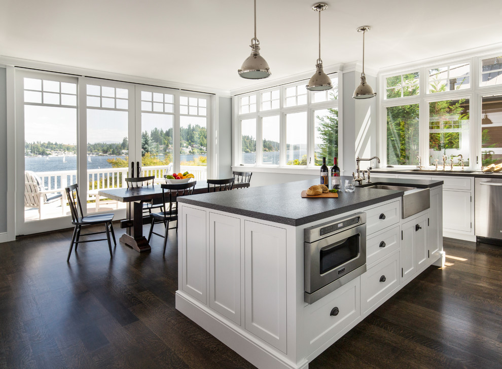 Mid-sized arts and crafts eat-in kitchen in Seattle with a farmhouse sink, shaker cabinets, white cabinets, window splashback, stainless steel appliances, dark hardwood floors, with island and brown floor.