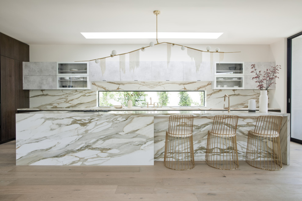 This is an example of an expansive contemporary l-shaped kitchen in Los Angeles with a drop-in sink, flat-panel cabinets, white splashback, stone slab splashback, stainless steel appliances, light hardwood floors, with island, brown floor and white benchtop.