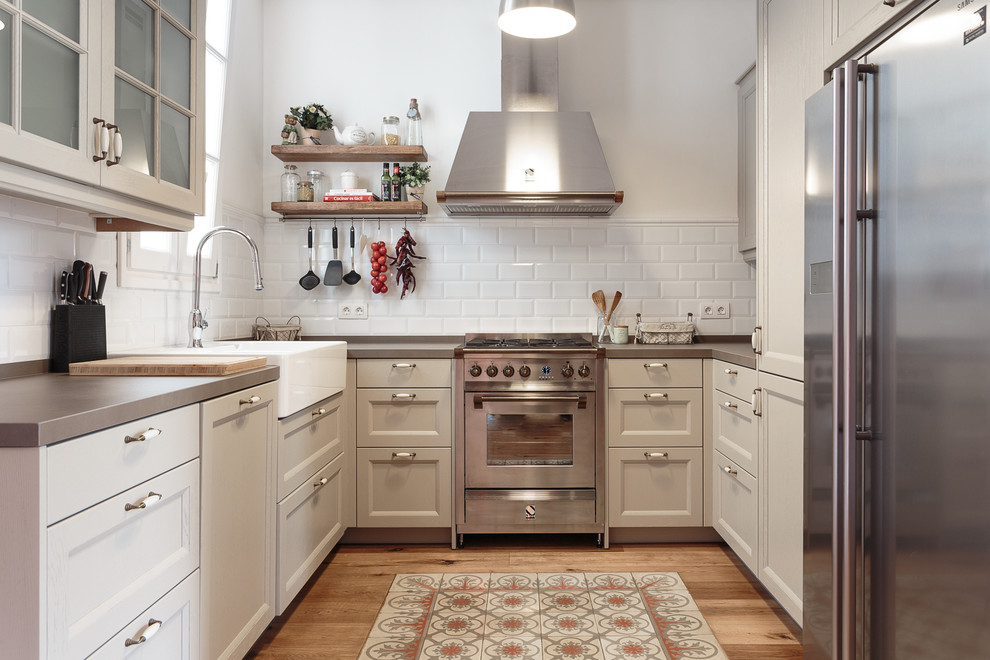 Inspiration for a mid-sized transitional u-shaped kitchen in Barcelona with recessed-panel cabinets, beige cabinets, white splashback, subway tile splashback, stainless steel appliances, no island, a farmhouse sink, light hardwood floors and grey benchtop.