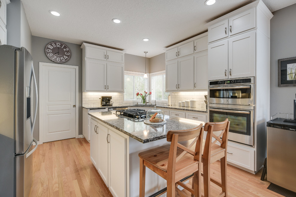 Example of a mid-sized transitional l-shaped light wood floor and brown floor enclosed kitchen design in Portland with a drop-in sink, shaker cabinets, white cabinets, quartz countertops, white backsplash, subway tile backsplash, an island and stainless steel appliances