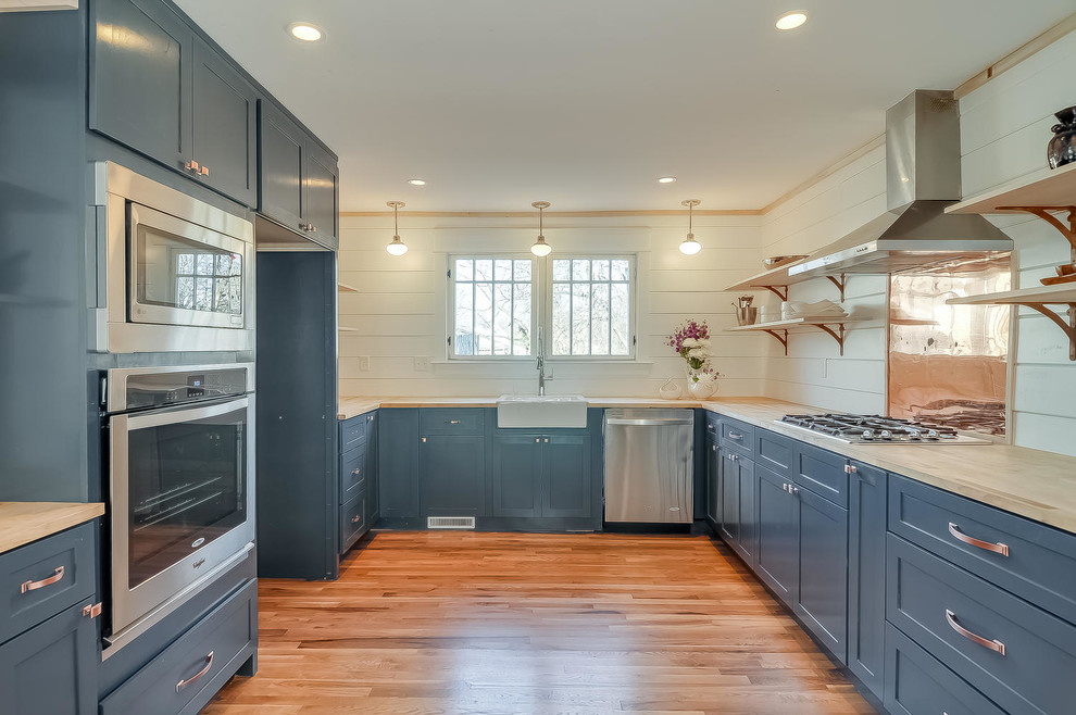 Mid-sized traditional u-shaped eat-in kitchen in Nashville with a farmhouse sink, shaker cabinets, blue cabinets, wood benchtops, white splashback, stainless steel appliances and light hardwood floors.