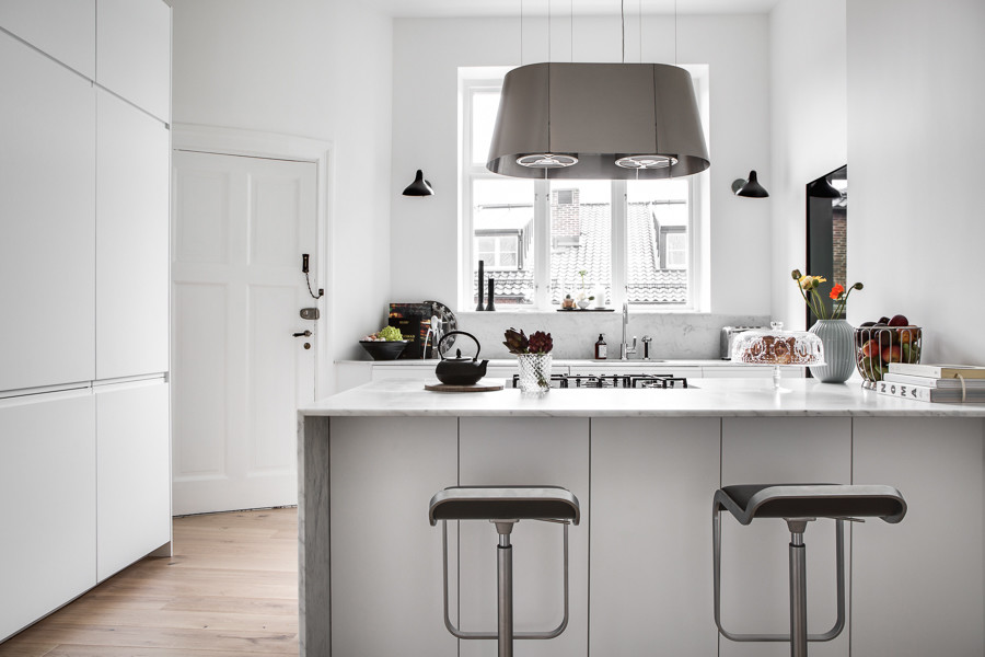 This is an example of a large scandinavian separate kitchen in London with an integrated sink, flat-panel cabinets, white cabinets, marble benchtops, white splashback, marble splashback, black appliances, light hardwood floors, with island and white benchtop.