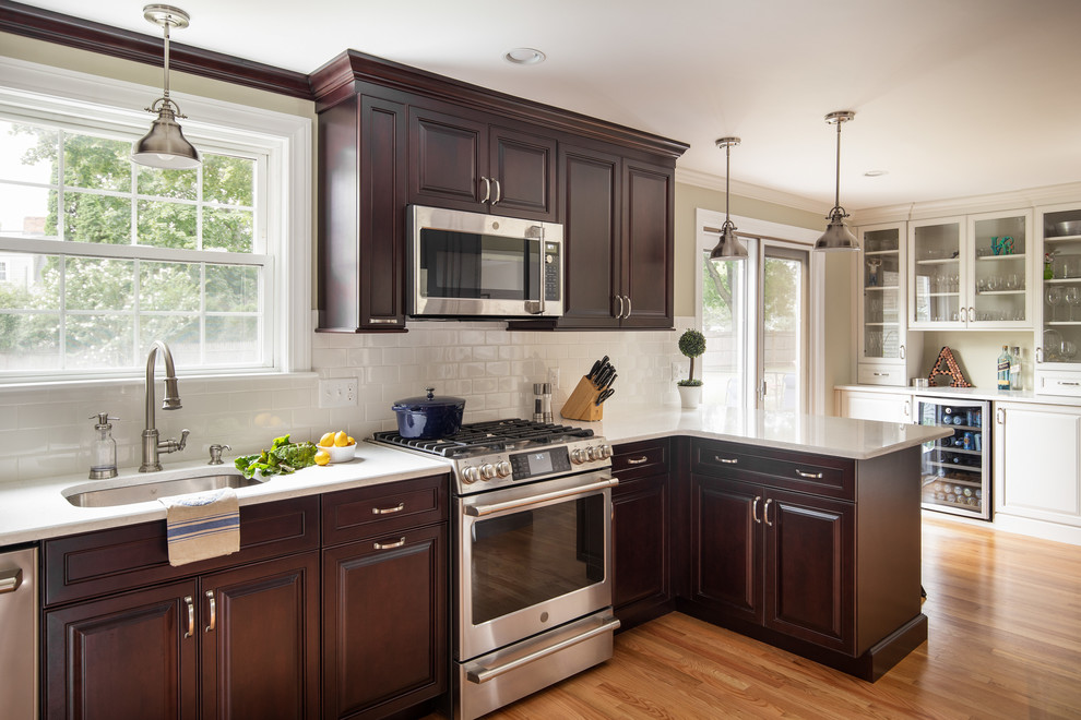 This is an example of a mid-sized transitional l-shaped eat-in kitchen in Boston with an undermount sink, raised-panel cabinets, dark wood cabinets, quartz benchtops, white splashback, ceramic splashback, stainless steel appliances, light hardwood floors, a peninsula and white benchtop.