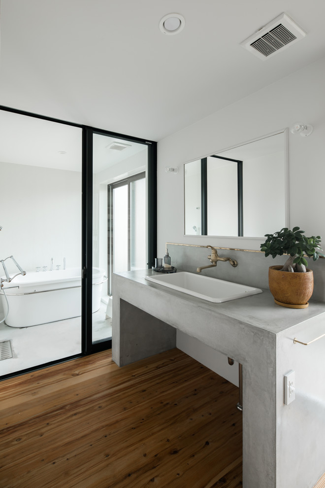 This is an example of a modern master bathroom in Other with a freestanding tub, white tile, concrete benchtops, white walls, medium hardwood floors, a drop-in sink, brown floor and grey benchtops.