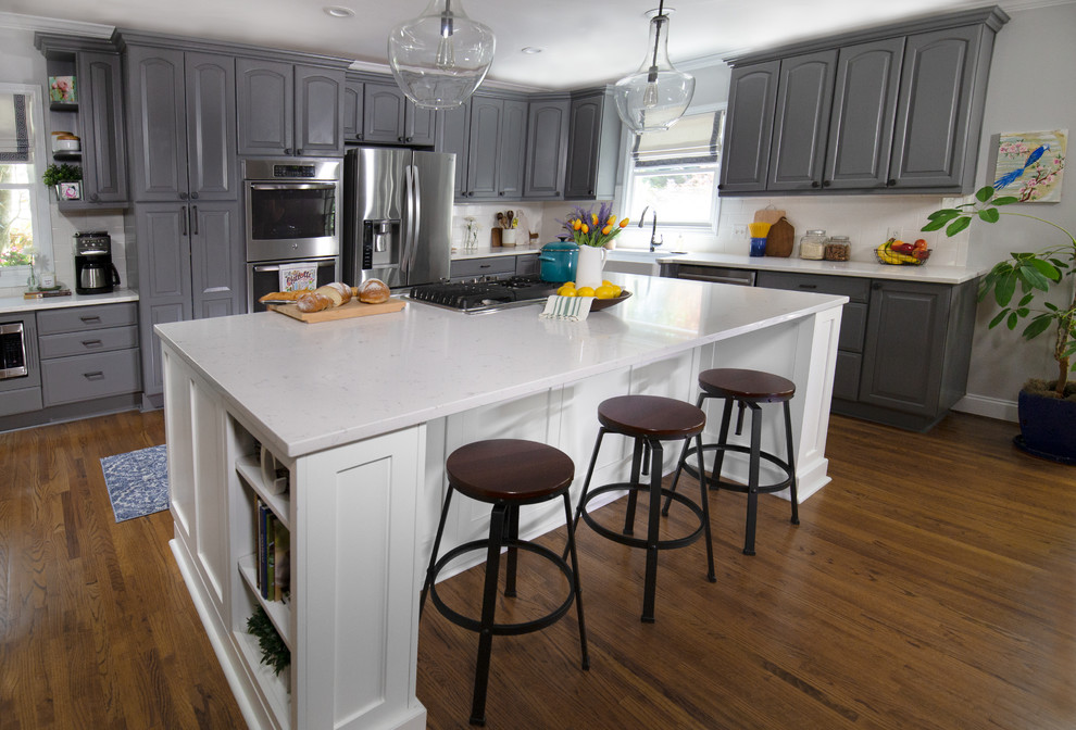 This is an example of a large country l-shaped open plan kitchen in Charlotte with a farmhouse sink, raised-panel cabinets, grey cabinets, quartz benchtops, white splashback, ceramic splashback, stainless steel appliances, medium hardwood floors, with island, brown floor and white benchtop.