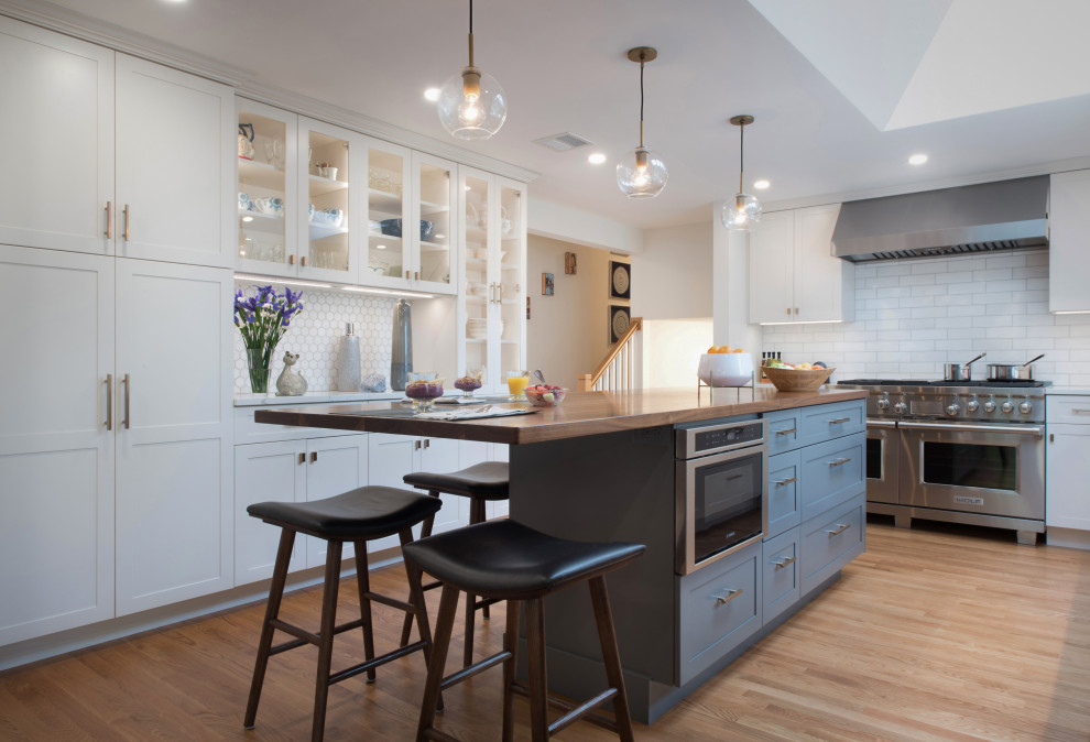 This is an example of a transitional u-shaped kitchen in Boston with shaker cabinets, white cabinets, white splashback, stainless steel appliances, light hardwood floors, with island, beige floor and white benchtop.