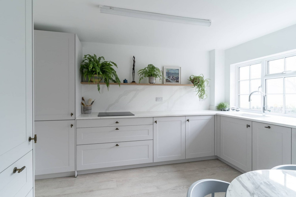 This is an example of a mid-sized traditional separate kitchen in Oxfordshire with a single-bowl sink, recessed-panel cabinets, white cabinets, quartzite benchtops, white splashback, black appliances, light hardwood floors, no island, beige floor and white benchtop.
