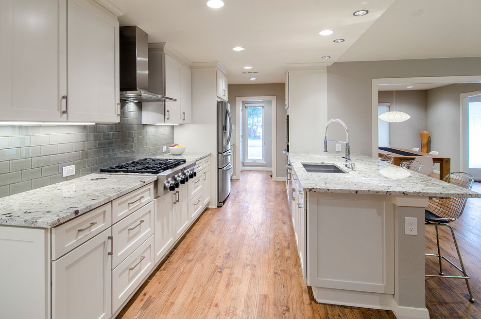 This is an example of a mid-sized contemporary single-wall open plan kitchen in Dallas with an undermount sink, shaker cabinets, white cabinets, granite benchtops, grey splashback, glass tile splashback, stainless steel appliances, light hardwood floors and with island.