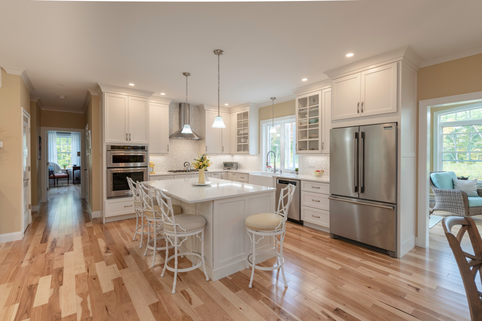 This is an example of a large transitional l-shaped eat-in kitchen in Boston with a farmhouse sink, shaker cabinets, white cabinets, quartz benchtops, white splashback, mosaic tile splashback, stainless steel appliances, medium hardwood floors, with island, brown floor and white benchtop.