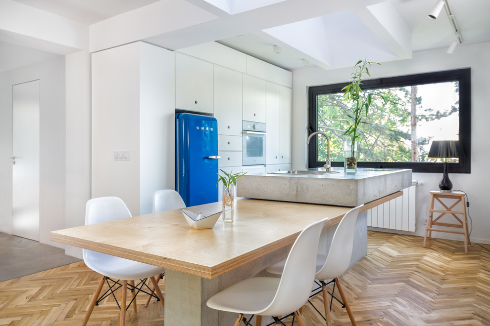 Photo of a contemporary kitchen in Other with flat-panel cabinets, white cabinets, coloured appliances, light hardwood floors, with island and concrete benchtops.