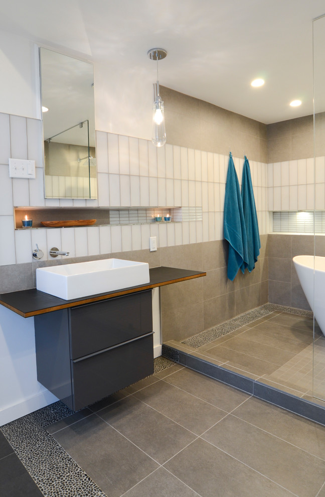 Photo of a large modern master wet room bathroom in Raleigh with flat-panel cabinets, grey cabinets, a freestanding tub, a wall-mount toilet, gray tile, ceramic tile, white walls, ceramic floors, a vessel sink, grey floor, an open shower and wood benchtops.