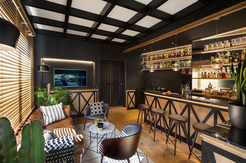 Design ideas for a transitional galley seated home bar in Barcelona with raised-panel cabinets, beige cabinets, mirror splashback, medium hardwood floors, brown floor and black benchtop.