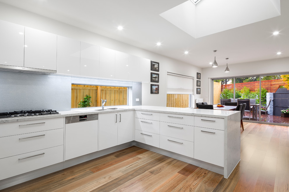 Design ideas for a mid-sized modern u-shaped open plan kitchen in Melbourne with a double-bowl sink, flat-panel cabinets, white cabinets, marble benchtops, white splashback, metal splashback, white appliances, light hardwood floors and no island.